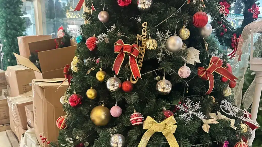 Un árbol de Navidad bellamente decorado con diversos adornos en colores rojo, dorado y plateado. El árbol está adornado con cintas, adornos y copos de nieve, colocado en el interior con cajas al fondo.