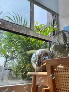 Una habitación iluminada por el sol, con una ventana desde la que se ve una exuberante vegetación, una brillante bola de discoteca colocada sobre una silla y un ventilador metálico. Las burbujas flotan en el aire y contribuyen a crear un ambiente sereno.