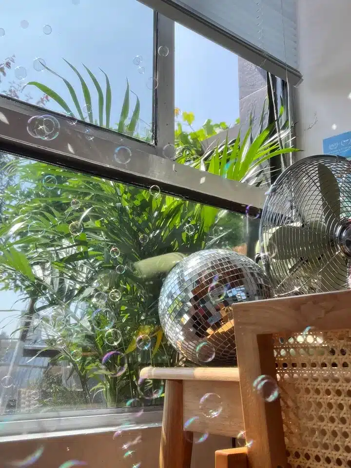 A sunlit room featuring a window with lush greenery visible outside, a shiny disco ball placed on a chair, and a metallic fan. Bubbles float through the air, adding to the serene atmosphere.