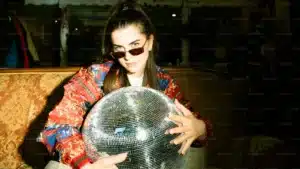 Person wearing sunglasses and a colorful jacket holding a large disco ball