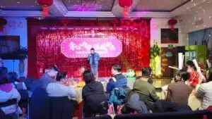A man standing on a stage with a pink glittering backdrop, addressing an audience seated at round tables decorated for an event.