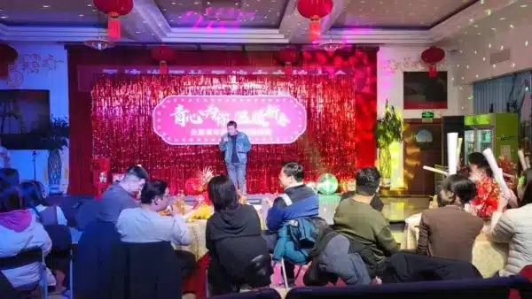 Un hombre de pie en un escenario con un fondo de purpurina rosa, dirigiéndose a un público sentado en mesas redondas decoradas para un evento.