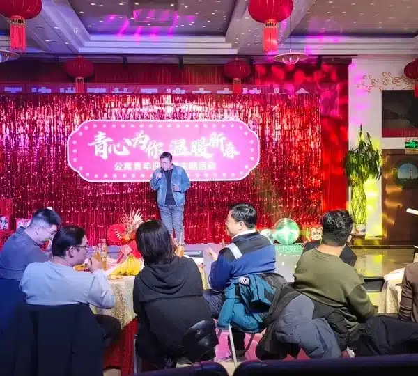 Un hombre de pie en un escenario con un fondo de purpurina rosa, dirigiéndose a un público sentado en mesas redondas decoradas para un evento.