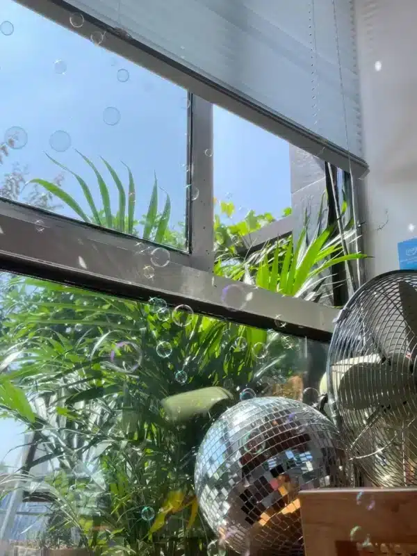 A bright room with a large window showing outdoor greenery and blue sky, featuring bubbles, a disco ball, and a fan.