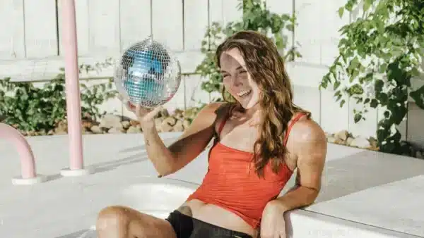 Mujer en bañador rojo sujetando una bola de discoteca sentada junto a una piscina.
